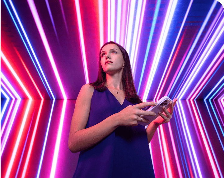 Jovem mulher com cabelo castanho escuro usando vestido azul, segurando um celular, diante de fundo com luzes neon coloridas, ideal para tecnologia e inovação.