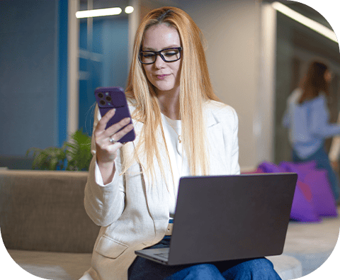 Mulher com cabelo ruivo usando óculos e vestida com blazer branco, sentada em ambiente de escritório moderno, olhando para celular enquanto trabalha com laptop.