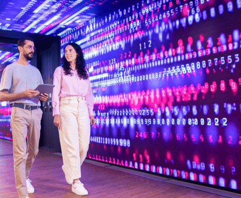Homens e uma mulher explorando uma parede de display digital com números e códigos coloridos brilhantes em uma sala moderna, destacando tecnologia e inovação.
