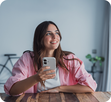 Mulher sorridente em ambiente interno, usando um smartphone cinza apoiado sobre a mesa de madeira.
