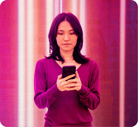 Mulher de cabelo escuro usando blusa roxa e segurando um smartphone, em fundo com luzes coloridas, representando o uso de tecnologia e conexão digital.