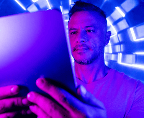 Homem usando tablet em ambiente com iluminação azul e roxa, expressão de concentração e foco na tela, cenário futurista ou tecnológico.