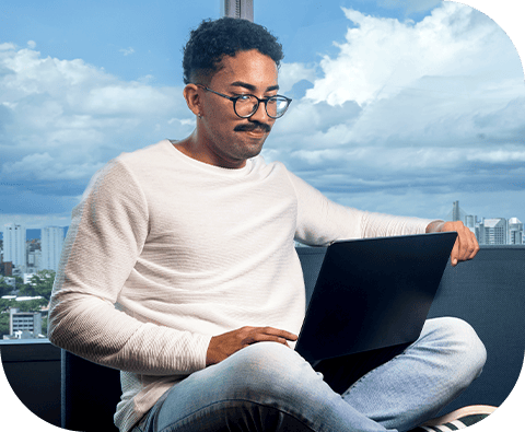 Homem usando óculos e suéter claro, sentado em ambiente com vista urbana, trabalhando com laptop.