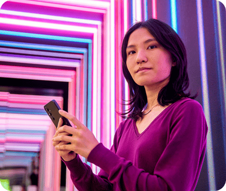 Mulher jovem com cabelo castanho escuro segurando um smartphone, vestindo blusa roxa, em corredor com iluminação de neon colorido em tons de azul, rosa e roxo.