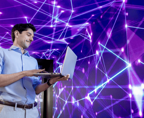 Homem sorridente segurando um laptop, com fundo de luzes e conexões digitais demonstrando tecnologia e inovação.
