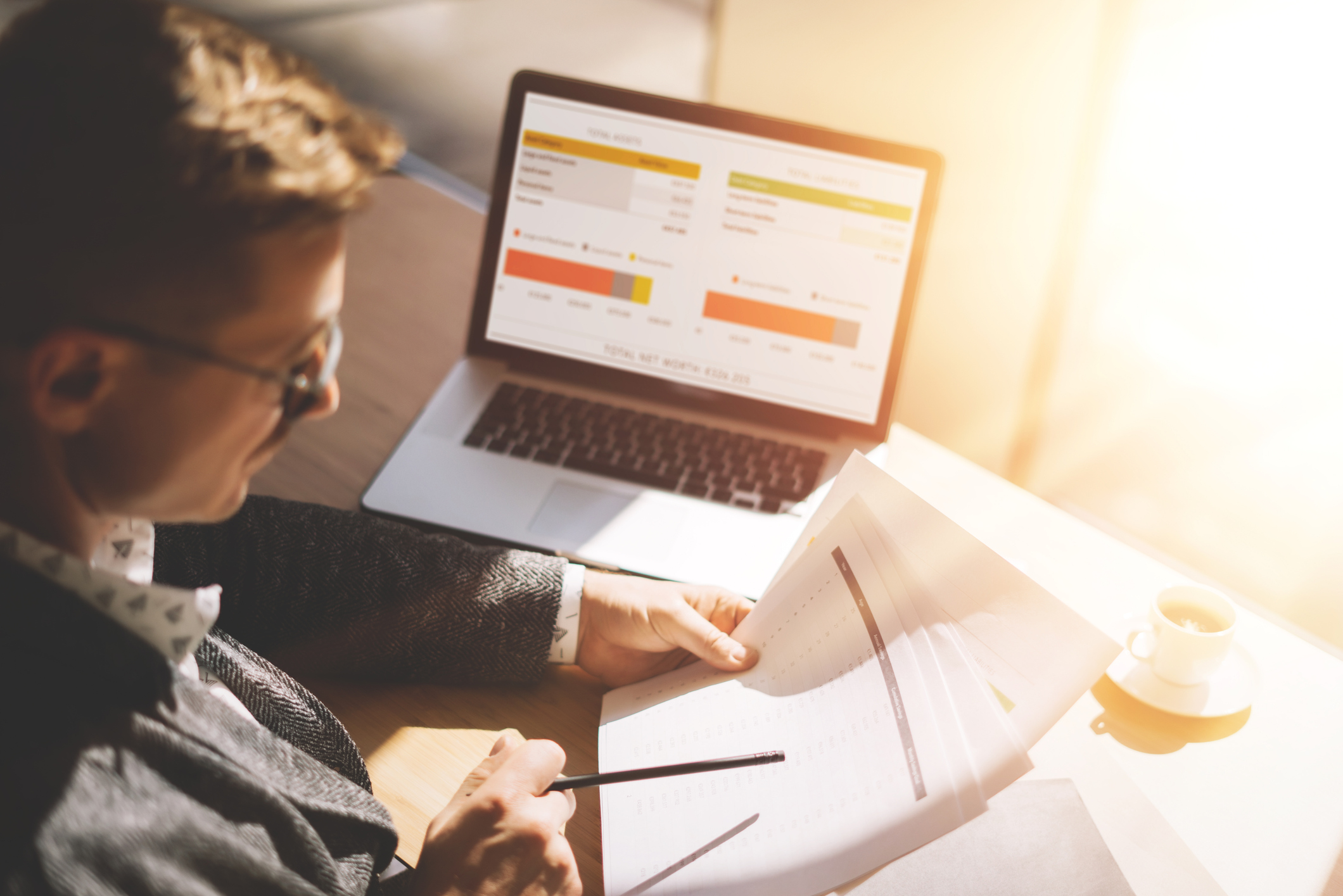 Adult businessman in eyeglasses working at sunny office on laptop while sitting at the wooden table.Man analyze document in his hands.Graphs and diagramm on notebook screen.Blurred background