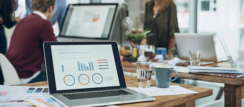 Closeup shot of a laptop with graphs on the screen and a team working in the background