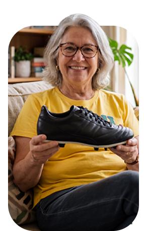 Mulher idosa sorridente segurando um tênis preto de futebol, sentada em ambiente aconchegante com plantas na decoração.
