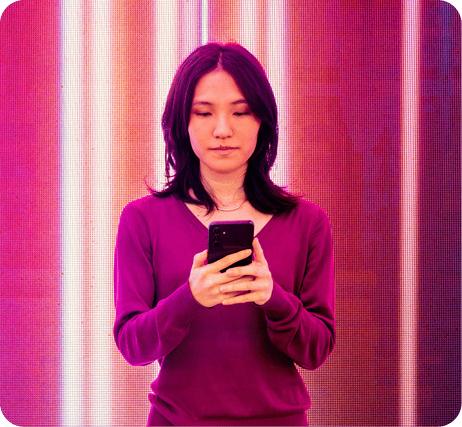 Mulher de cabelo escuro usando blusa roxa e segurando um smartphone, em fundo com luzes coloridas, representando o uso de tecnologia e conexão digital.