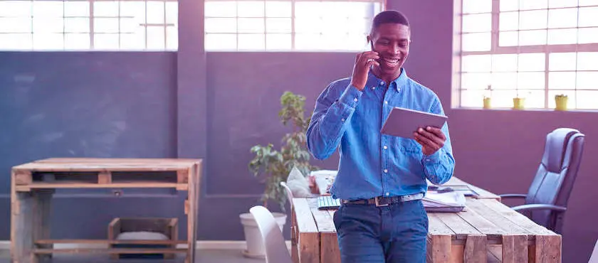 A imagem mostra um homem negro, sorrindo e falando ao celular com a mão direita e olhando algumas informa~ções no tablet com a mão esquerda. Ele usa uma camisa jeans azul claro e usa uma calça jeans escura e está apoiado em uma mesa feita de pallets. A imagem dá a entender que o homem está analisando como fazer a expansão do seu negócio.